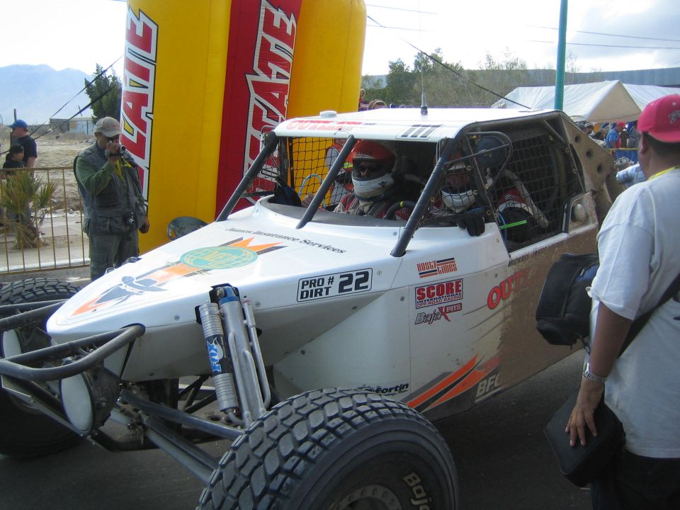 White and orange off-road buggy number 22 with two helmeted occupants near Tecate arch
