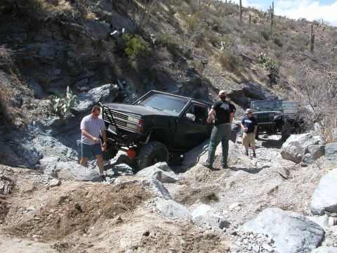 1984 Ford Bronco - Two off-road vehicles are navigating a difficult, rocky trail in a desert environment.