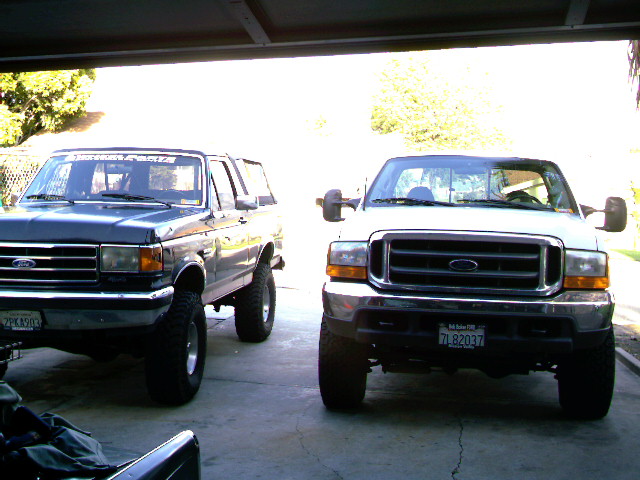 White Ford Super Duty and blue lifted Bronco parked side by side under a carport