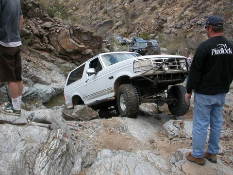 1984 Ford Bronco - Modified white Ford Bronco tackling a difficult rocky trail with other vehicles in the distance.