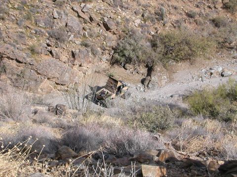1984 Ford Bronco - A damaged vehicle rests off-road in a dry, rocky environment, partially obscured by desert brush.