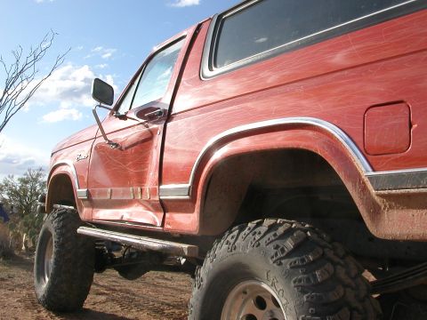 1984 Ford Bronco - Side view of a red Ford Bronco, lifted with off-road tires, on a dirt road.