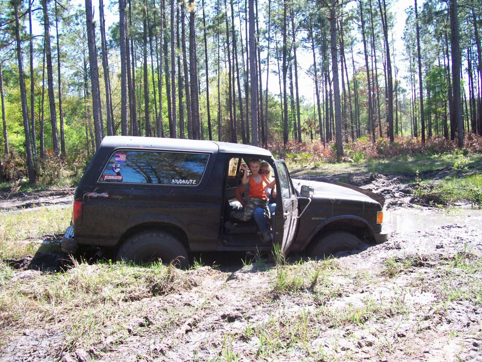 Mud-covered Ford Bronco stuck in a wet rut, person seated in open doorway