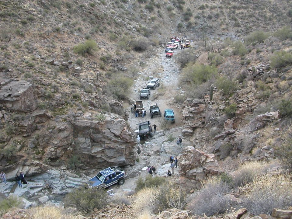 Several off-road vehicles driving down a dry, rocky creek bed in a desert canyon
