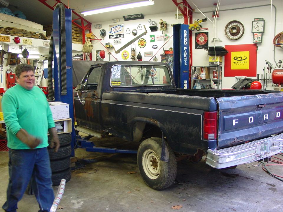 Man in green shirt stands next to dark blue Ford pickup on a lift in a garage. Truck has 'FORD' on tailgate
