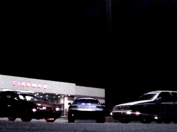 Three cars parked at night in front of a cinema with a lit red CINEMAS sign