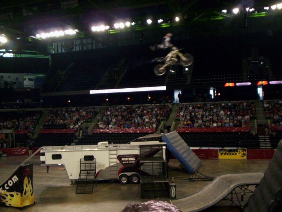 FMX rider hanging off back of white dirt bike mid-air over arena and GasGas trailer