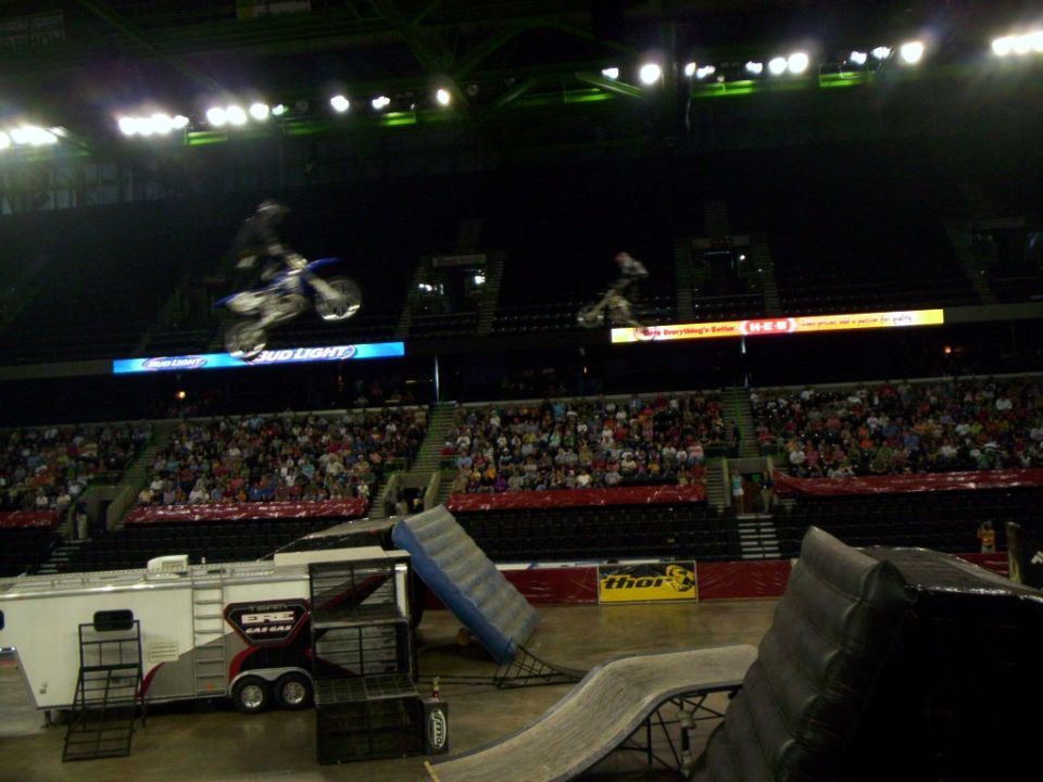 Two motocross riders in mid-air over a ramp setup inside an arena with spectators