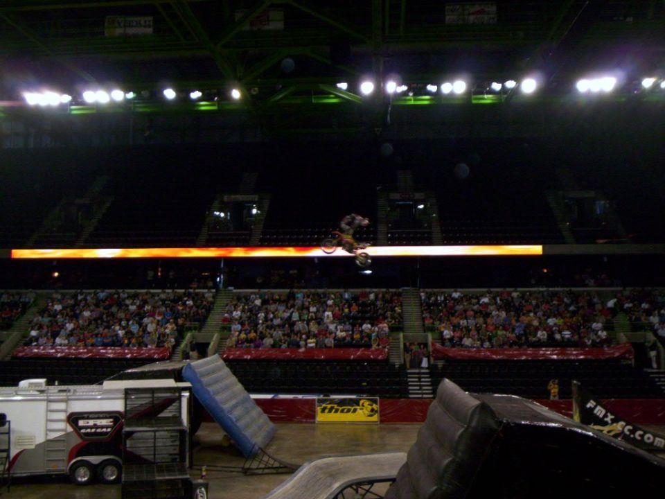 Motocross rider doing a trick mid-air over a crowd in an arena. Ramps and a trailer are visible