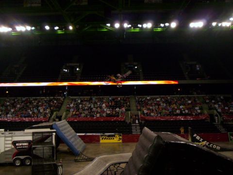 1986 Ford Bronco - Motocross rider mid-trick above an arena floor, body thrown off the seat with the bike sideways in front of a flame-graphic LED ribbon boar…