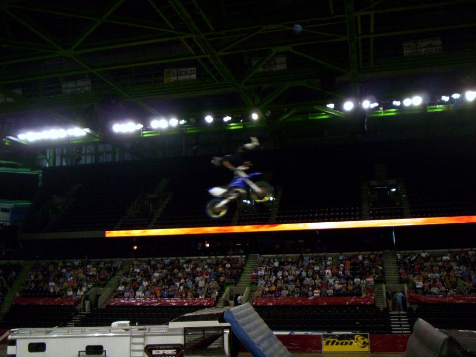 FMX rider extended superman-style above blue dirt bike near arena roof trusses