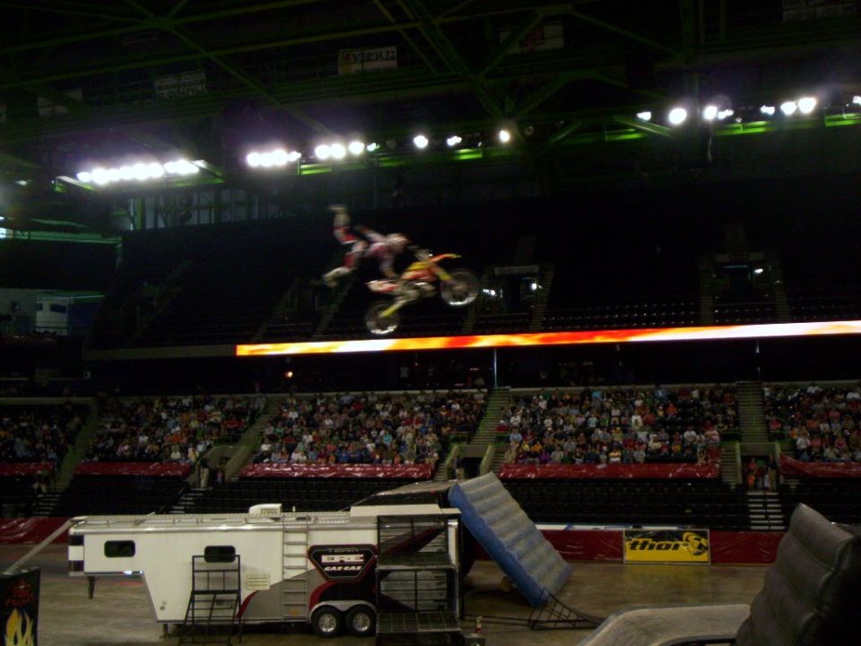 Rider hanging off a red dirt bike mid-jump above an arena crowd with race trailer below