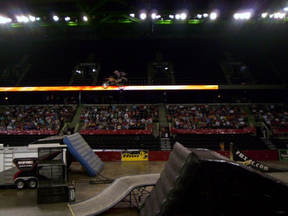 Motocross rider airborne over ramps inside a stadium with spectators