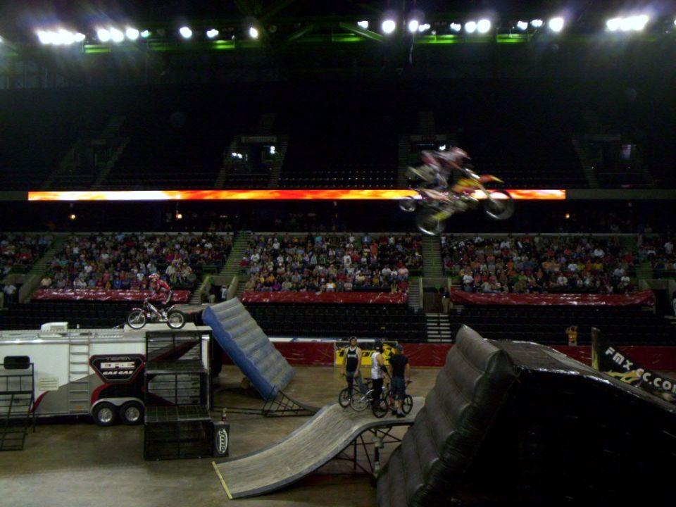 Motocross riders in mid-air during a stunt show inside an arena with a large crowd watching