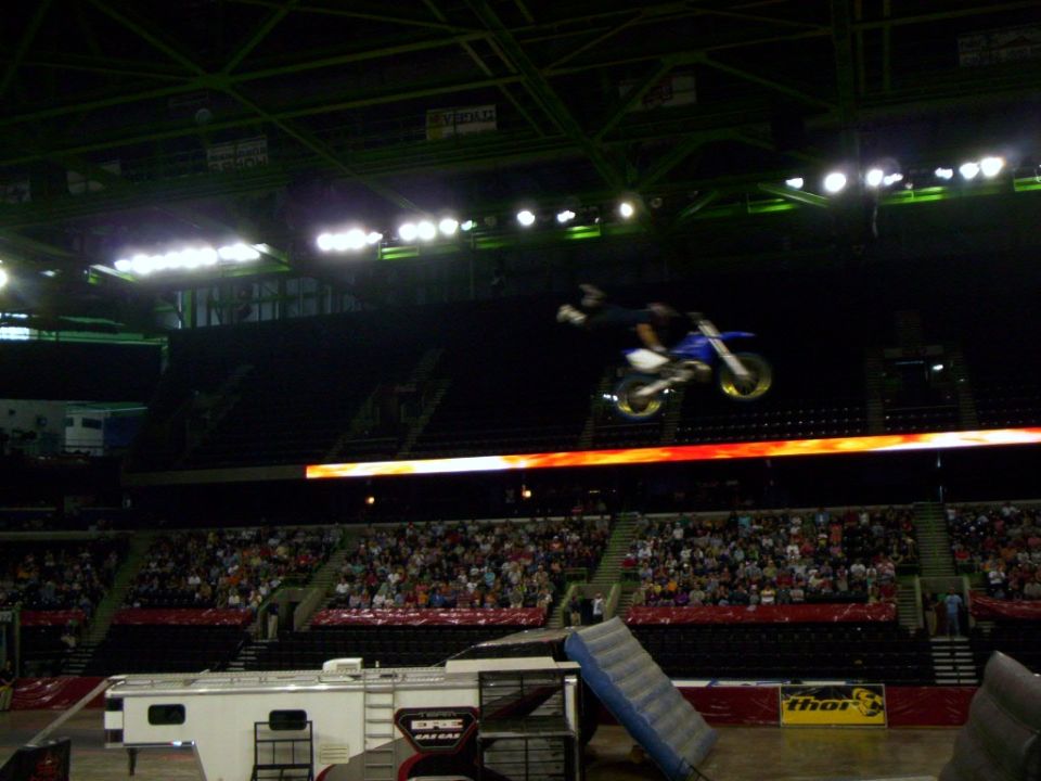 Rider extended off a blue dirt bike with yellow rims mid-air above an arena crowd