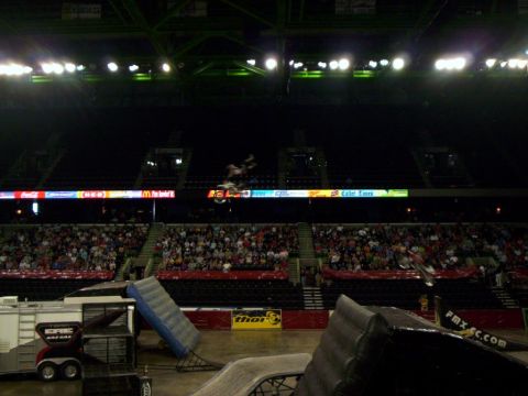 1986 Ford Bronco - Freestyle motocross rider mid-backflip high above an indoor arena floor, silhouetted against dark upper seating with a lit ring of sponsor…