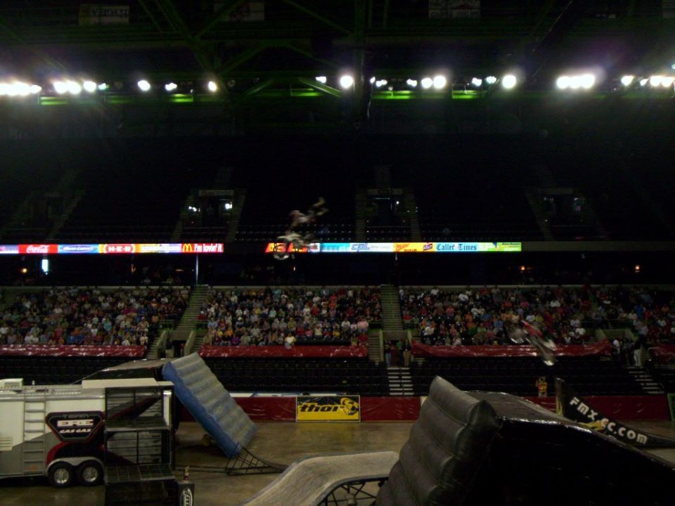 FMX rider mid-backflip over indoor arena ramps with sponsor banners and crowd