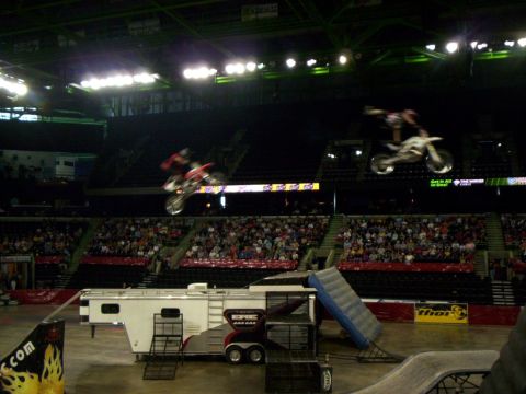 1986 Ford Bronco - Two freestyle motocross riders airborne at once inside an arena, one on a red bike doing a heel-clicker at left and one on a white bike ext…