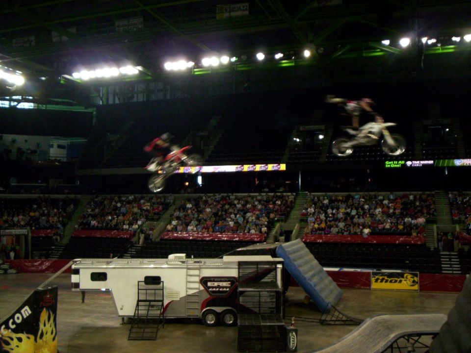 Two FMX riders airborne over arena floor with GasGas team trailer and ramps below