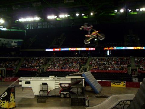 1986 Ford Bronco - Freestyle motocross rider laid out flat above a red and yellow dirt bike in a heel-clicker-style trick, airborne over an indoor arena. A wh…