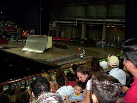 1986 Ford Bronco - View from the arena stands over spectators' heads to a rider in red riding a wheelie on a small trials-style bike across the concrete floor…