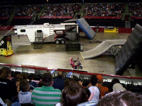 1986 Ford Bronco - Elevated view across an arena floor where a rider in red gear stands on a trials bike near the barrier as spectators watch. A white Team Ga…