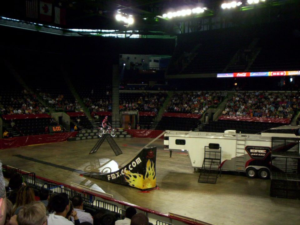 Motocross rider on a ramp inside an arena with spectators. FMX.com logo visible on ramp