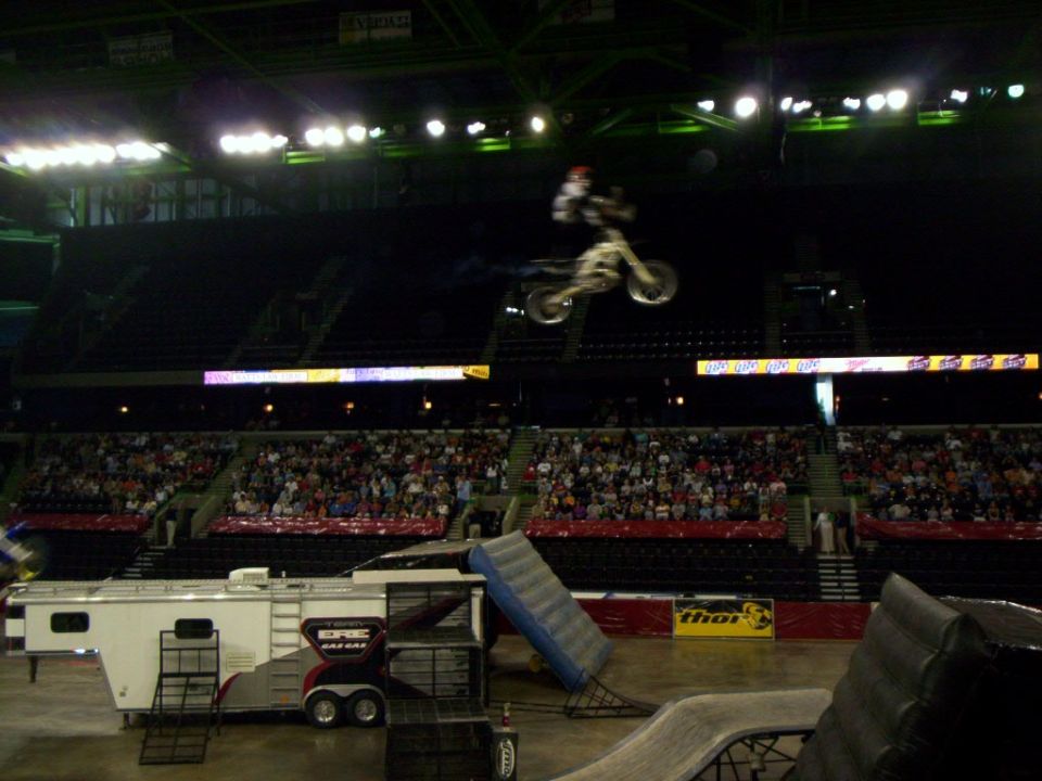 FMX rider extended above white dirt bike mid-trick over arena with GasGas trailer