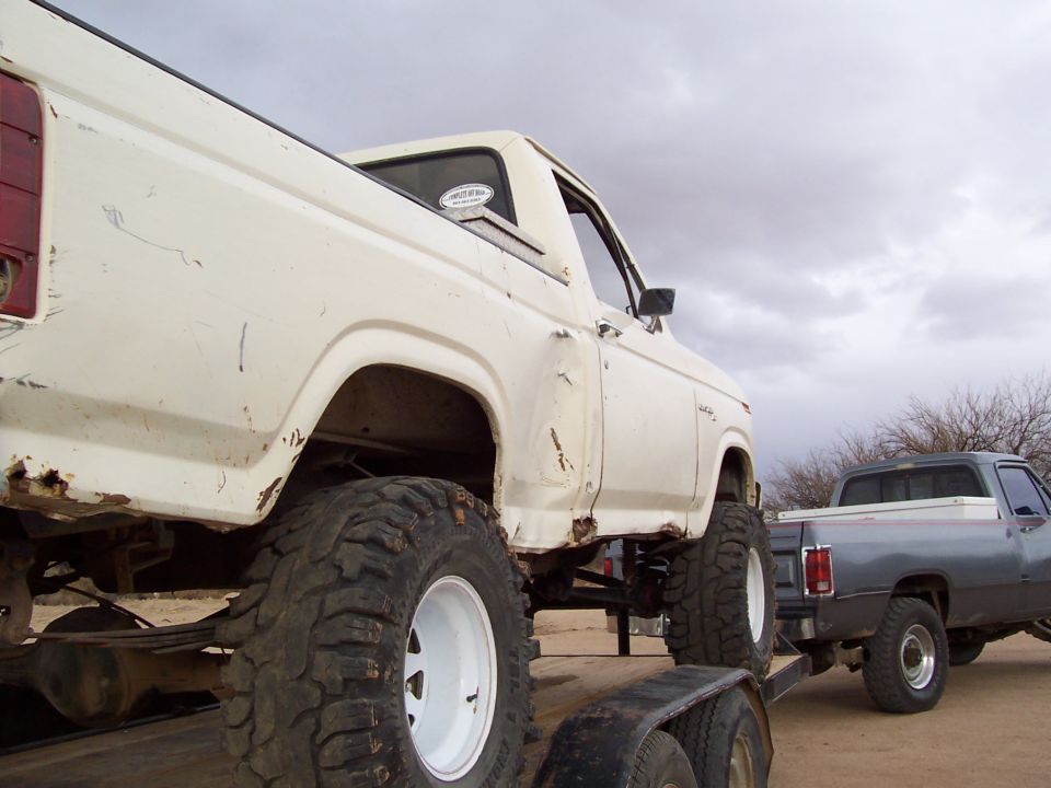 White Ford pickup with large tires on a trailer, next to a gray pickup truck
