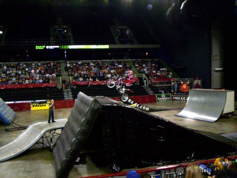 1986 Ford Bronco - Rider in red gear balances a trials bike on its rear wheel atop a black tarp-covered obstacle on an arena floor, with a spotter standing at…