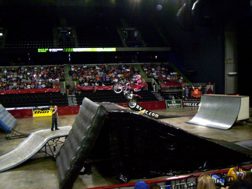 Trials rider wheelieing over a black obstacle on an arena floor with ramps and crowd