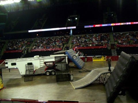 1986 Ford Bronco - Rider in red gear rides a trials bike up the face of a steep blue ramp on an arena floor while an announcer with a microphone stands on the…