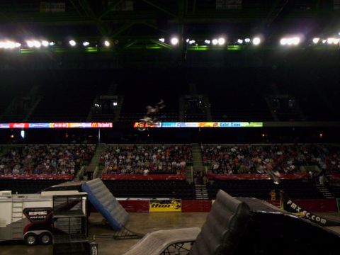 1986 Ford Bronco - Wide arena shot of a freestyle motocross rider inverted mid-backflip above the LED ribbon board, with a second bike blurred at right. The f…