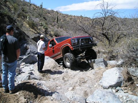 1993 Ford Bronco - A red 1993 Ford Bronco tackles a challenging rocky off-road path, with its front-right wheel significantly elevated.