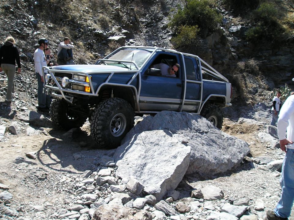 Blue pickup truck with custom roll cage and off-road tires navigating large rocks on a trail