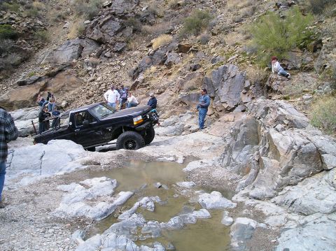 1993 Ford Bronco - A group of people watch as a modified truck attempts to drive through a rocky, water-filled obstacle course.