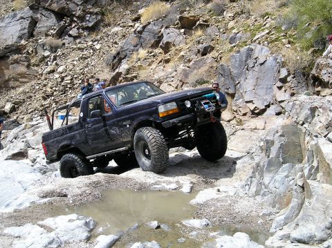 1993 Ford Bronco - A dark blue Ford Bronco tackles a challenging rocky trail with a puddle in the foreground.
