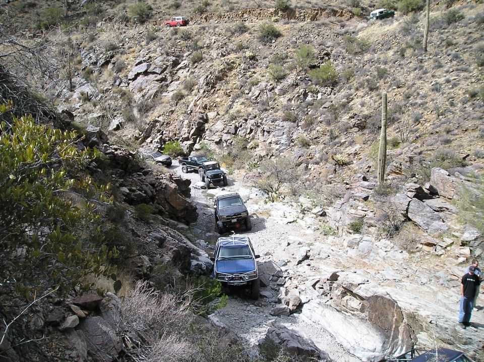 Multiple SUVs and trucks driving in single file on a rough, rocky desert trail