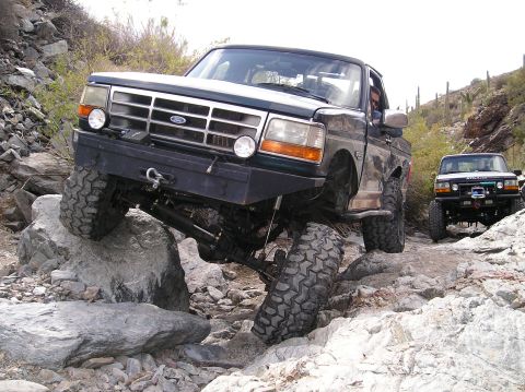 1993 Ford Bronco - Two Ford F-Series trucks navigate a rocky, off-road trail in a desert environment.