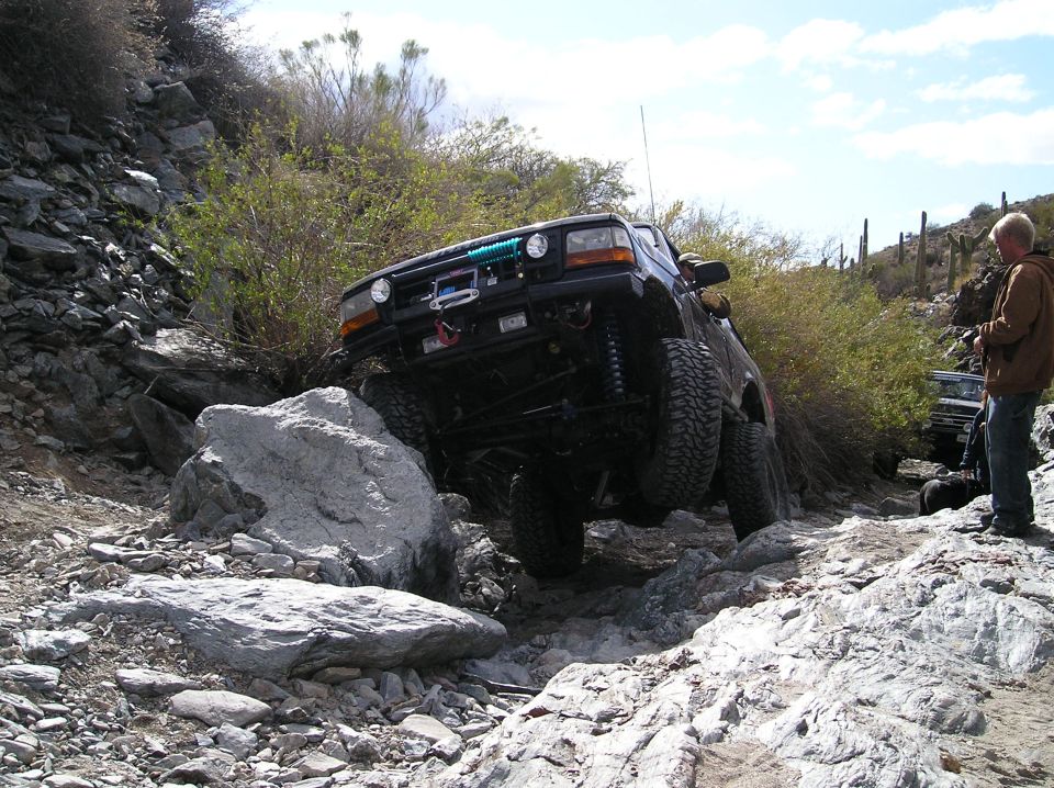 Dark blue Ford Bronco off-roading over rocks, front-left tire lifted
