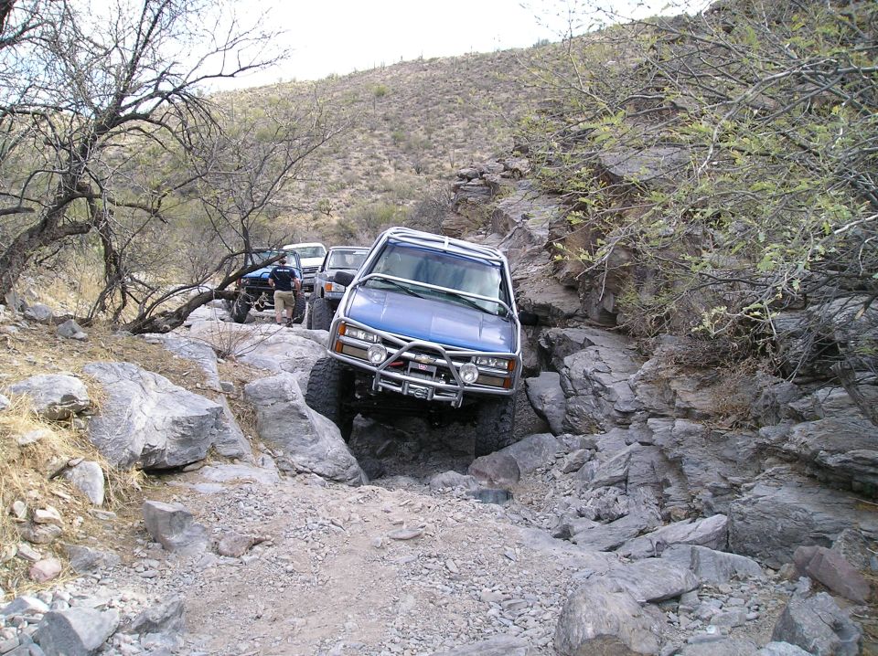 Blue SUV with aftermarket bumper and roll cage on a rocky trail, other off-road vehicles behind it
