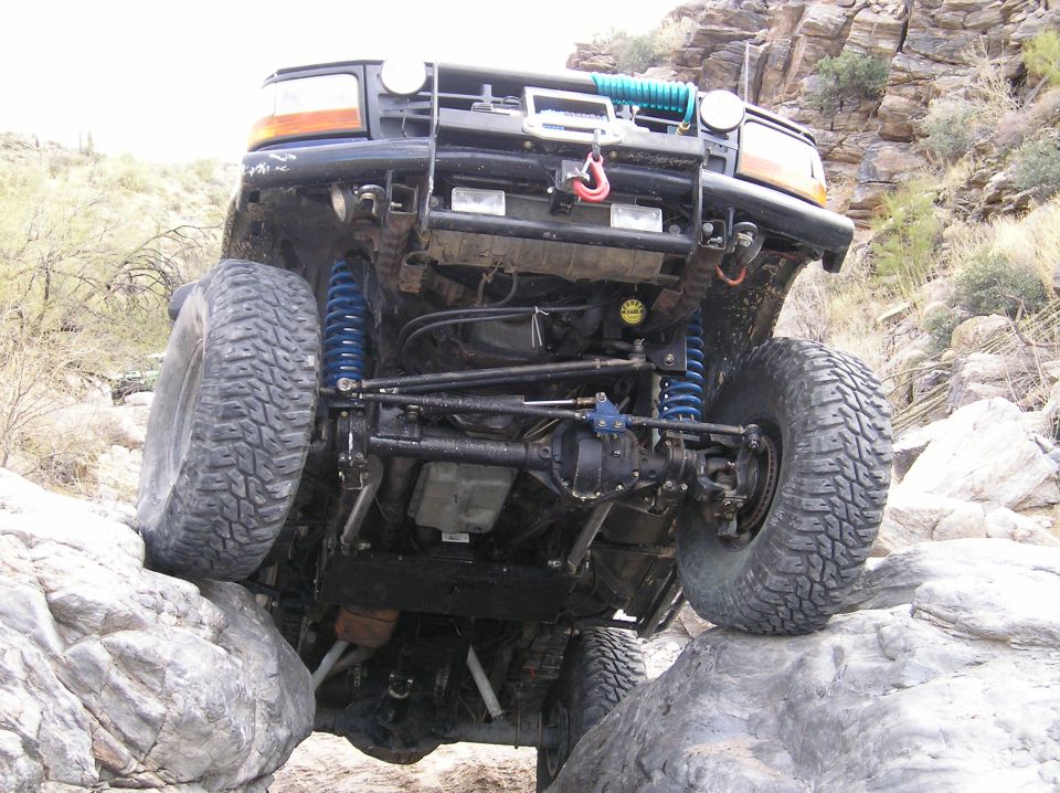Off-road vehicle with large tires climbing over rocks, showing undercarriage and front bumper with winch