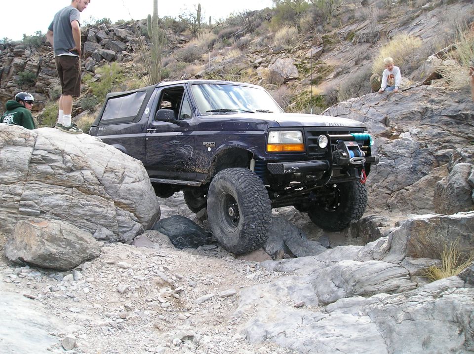 Dark blue 1993 Ford Bronco with large tires navigating a rocky desert trail, front winch visible