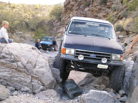 1993 Ford Bronco - A dark blue Ford F-Series truck is carefully driven over large rocks on an off-road trail.
