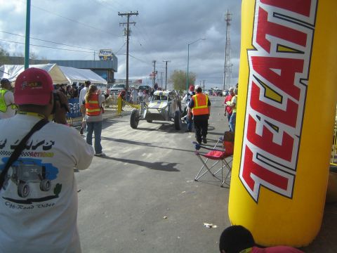 Off-road race buggy escorted by marshals down street with large yellow Tecate inflatable