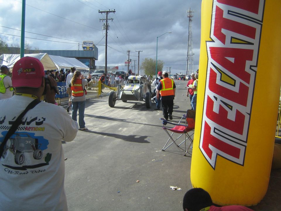 Off-road race buggy escorted by marshals down street with large yellow Tecate inflatable