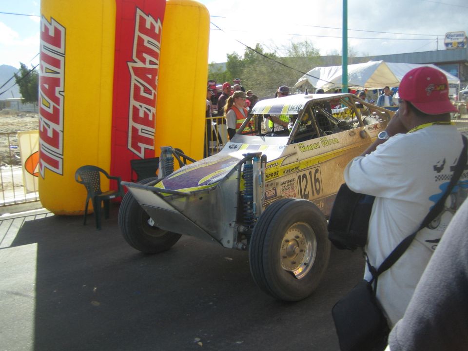 Yellow and purple off-road race buggy number 1216 beside Tecate arch with photographer