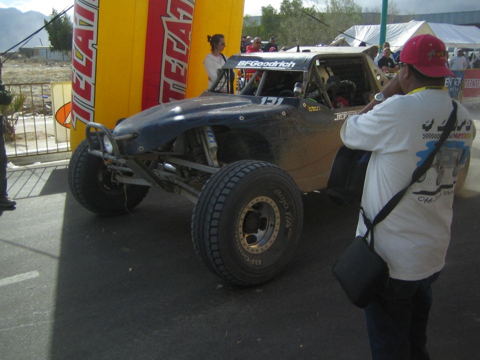 Dark off-road race buggy in paved staging area beside Tecate arch with photographer