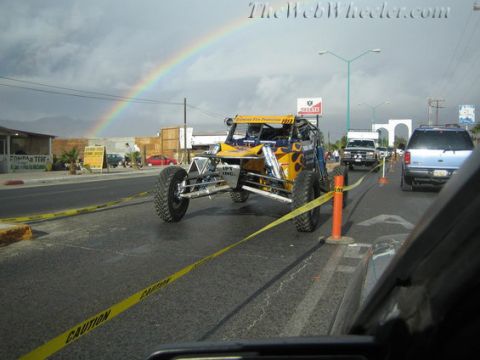 1990 Ford Bronco - yellow flame-painted off-road buggy sits on a street under a rainbow behind yellow caution tape, with traffic and shops in the background.