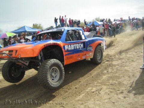 1990 Ford Bronco - orange and blue off-road race truck with FABTECH and Toyo Tires graphics climbs a dirt berm in front of a large lined-up crowd.